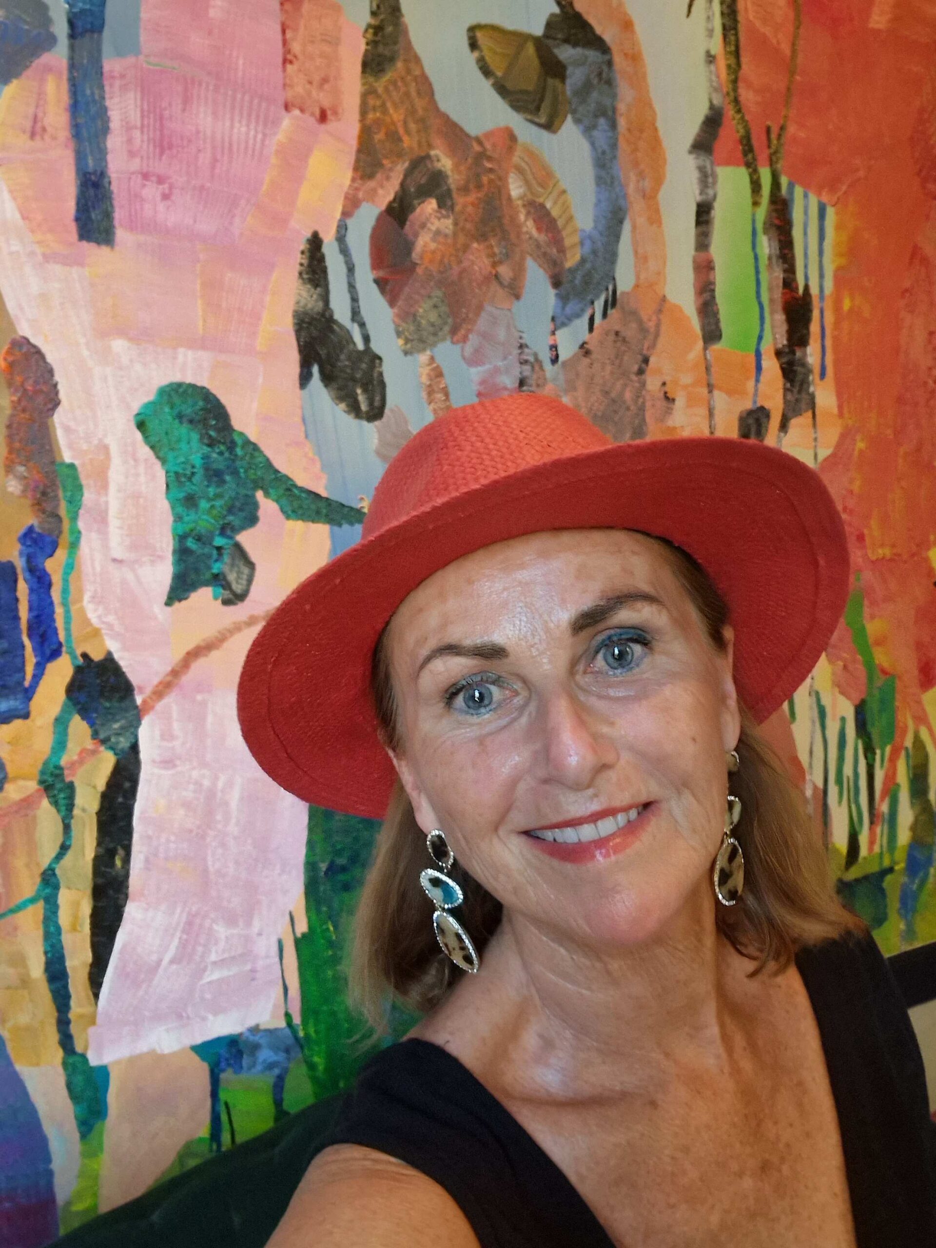 Wearing a red fedora in front of a wall mural, Nancy McGee, owner of Absolutely Southern France, smiles for a selfie.
