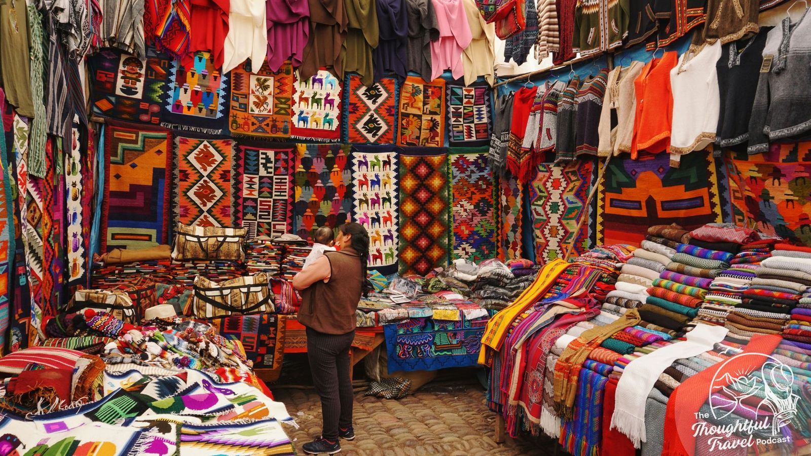 A woman holding a baby stands inside a market stall with the walls and tables covered in indigenous tapestry—Centre for Good Travel