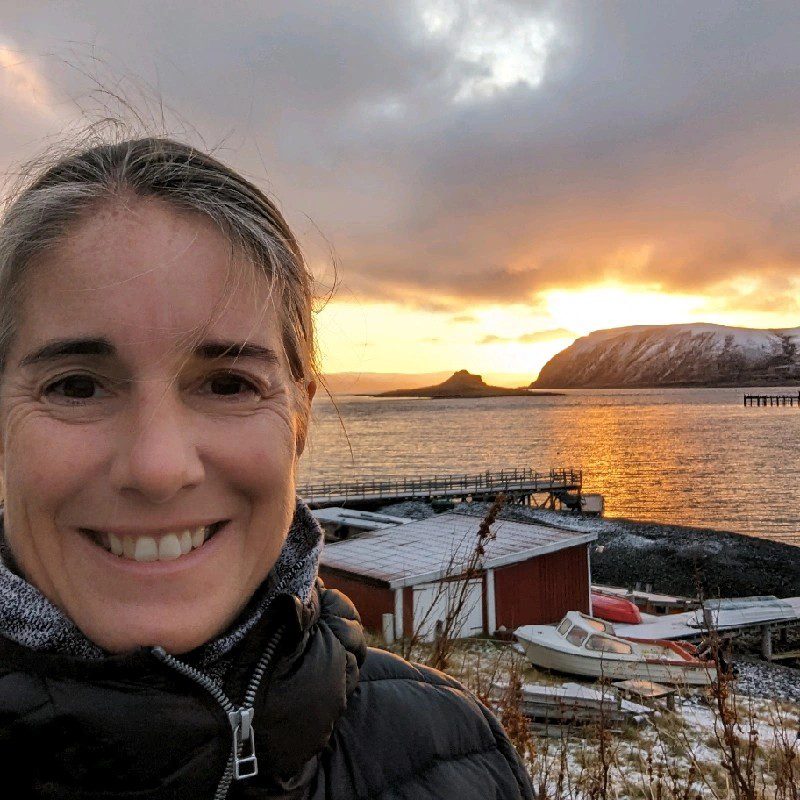 Carly Biggart, VP of Sales and Marketing for Hurtigruten Norway, smiles for a selfie with scenic fishing village and mountains in the background.
