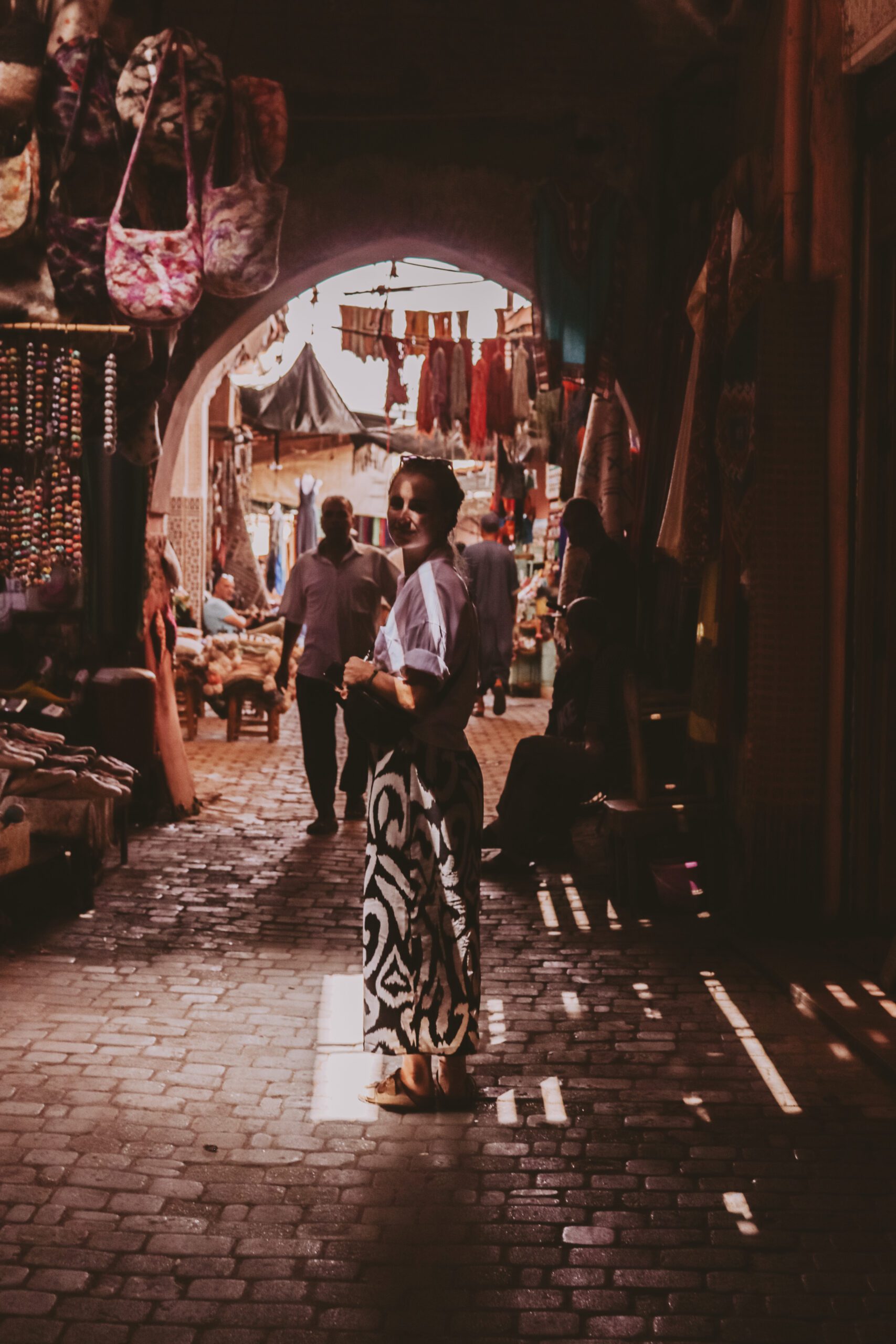 A woman standing in shadows in an alley in Morocco, wearing a long, patterned dress looks back at the camera. Sororal X Morocco