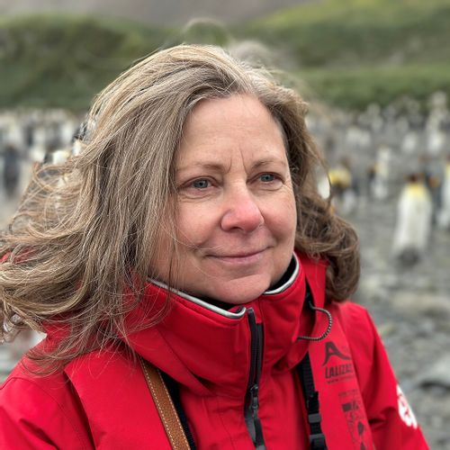 Marikra Roberson, co-founder of INCA, wears a red parka and smiles as emperor penguins frolic in the background
