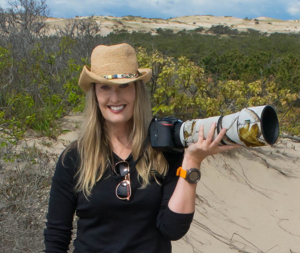 Sarah E Devlin, CEO and owner of Women in Wildlife Photography, poses for the camera wearing a straw cowboy hat and holding a long-lensed camera.