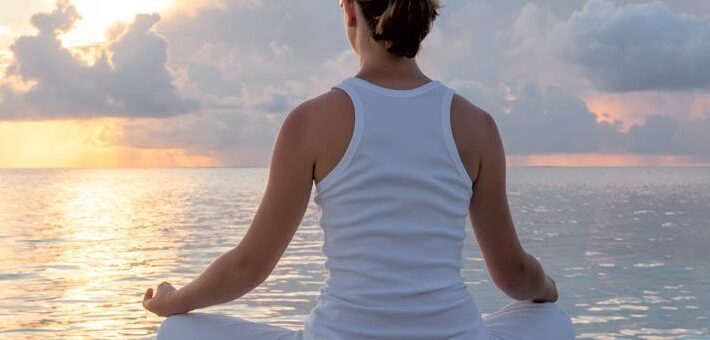 A young woman with her back to the camera sits in a meditative pose overlooking the ocean at sunset - Total Life Reset - Ultimate Retreat Company
