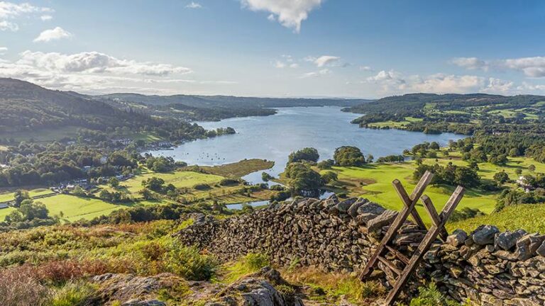View of Windermere in the English Lake District taken from Loughrigg Fell. On a summer morning. With white fluffy clouds, shafts of Light on to Green fields with a Dry Stone Wall and wooden Style in the Foreground.