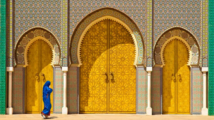 A woman wearing a striking blue burqa strides gracefully past the majestic golden doors of the historic Royal Palace of Fes - Best of Morocco, A Women-only Tour - Insight Vacations