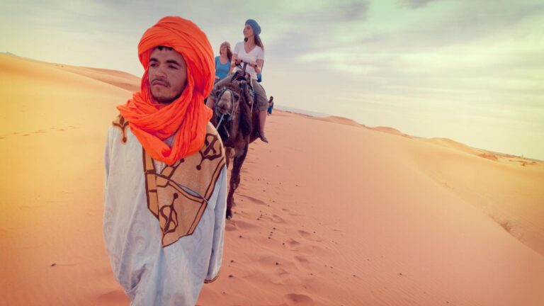 Man in a traditional outfit with a vibrant orange turban leads camels carrying tourists across vast desert sand dunes under a soft, cloudy sky.