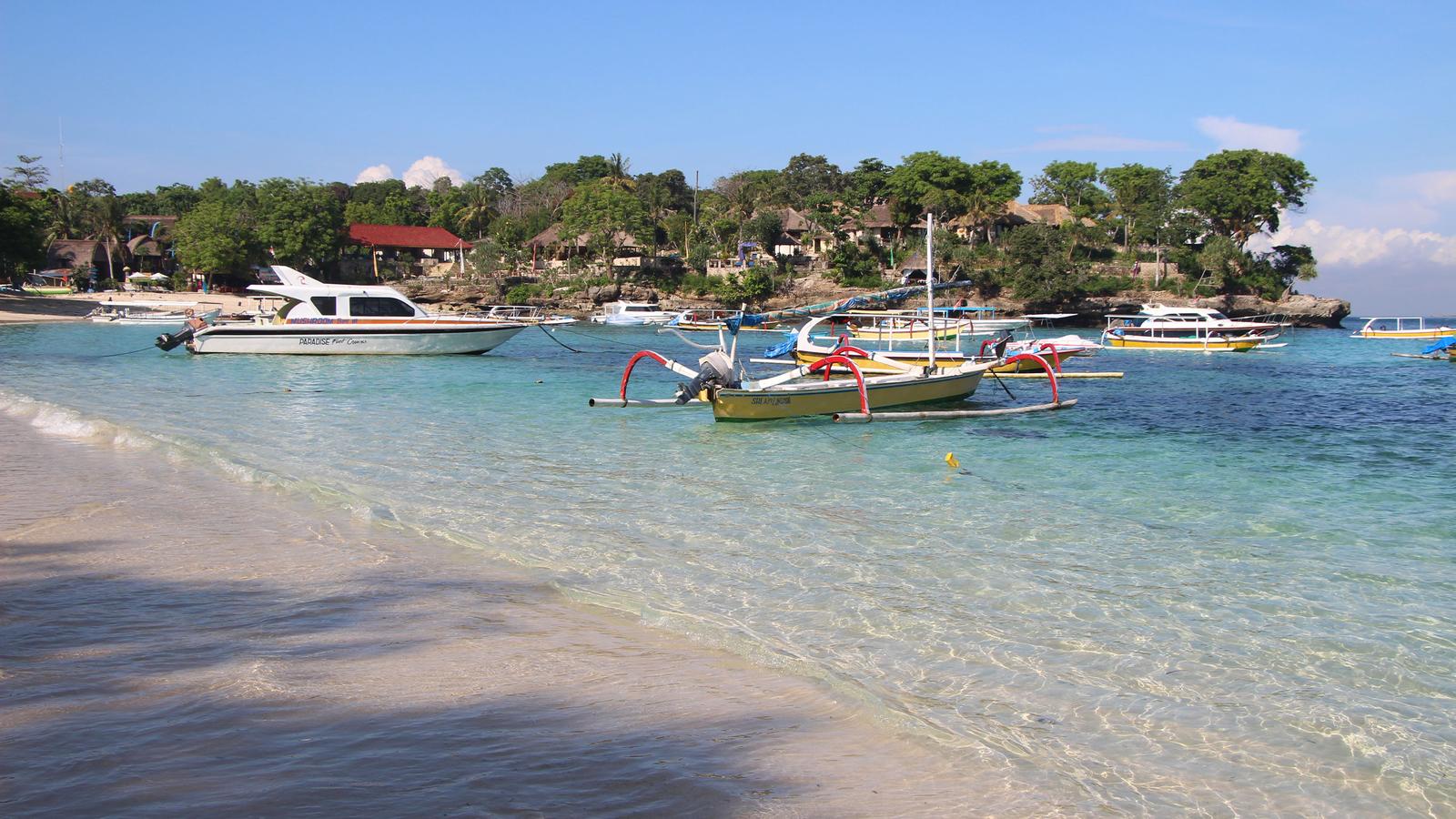 A scenic view of a beach in Bali with clear turquoise water, traditional fishing boats, and lush greenery in the background.