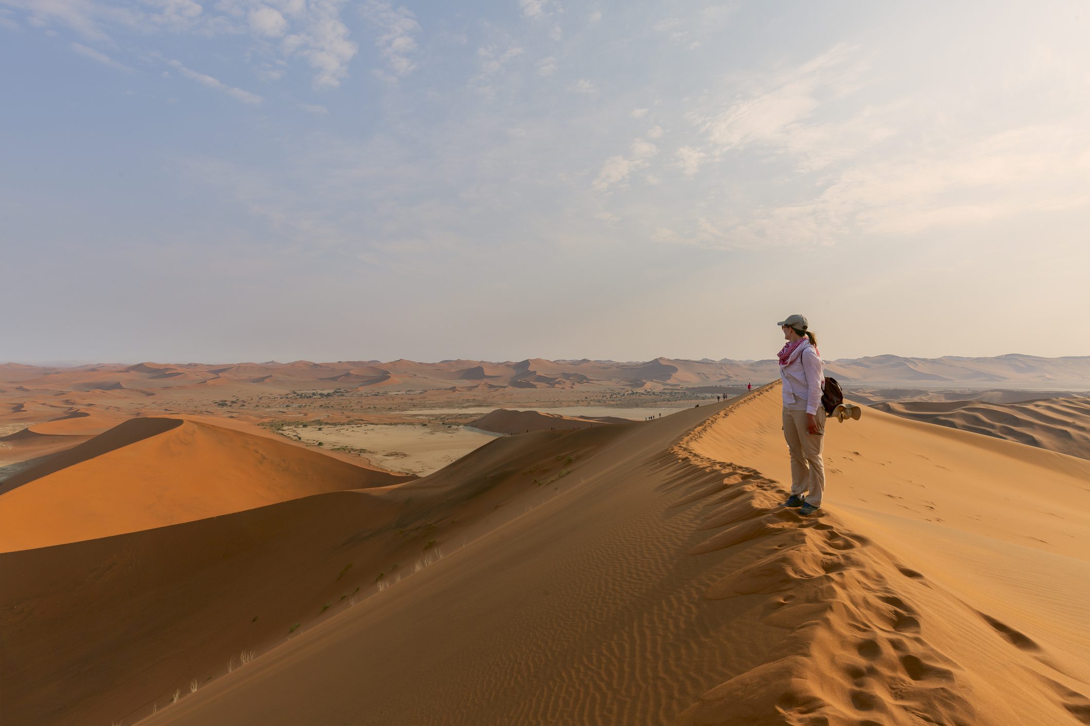 A person standing on top of a sand dune in a vast desert landscape, gazing into the distance under a partly cloudy sky.