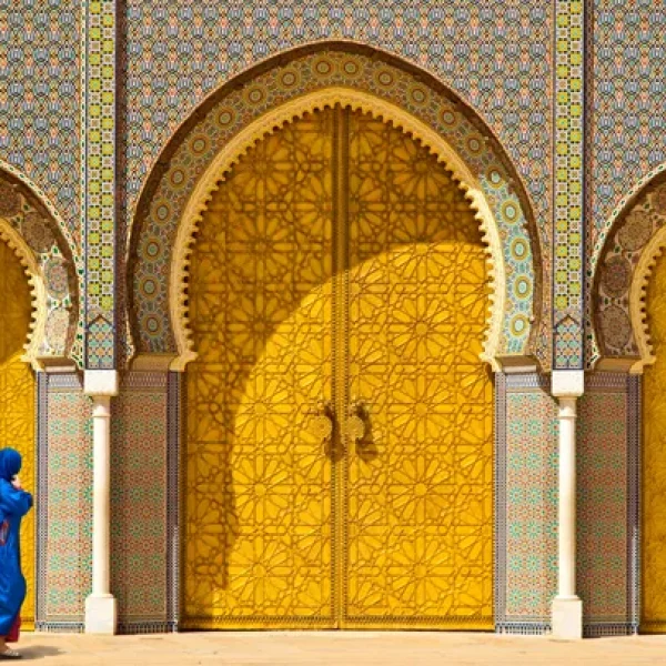 A woman wearing a striking blue burqa strides gracefully past the majestic golden doors of the historic Royal Palace of Fes - Best of Morocco, A Women-only Tour - Insight Vacations