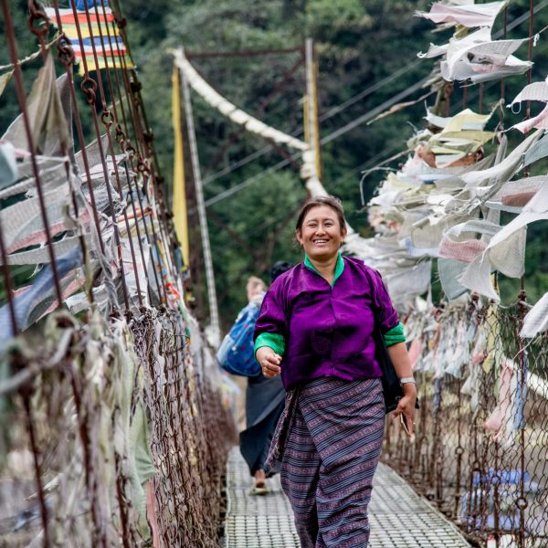 A Nepalese woman dressed in a colourful, cultural outfit, walks along a suspension bridge that has flags tied to the ropes to illustrate the Bhutan Hiking Adventure from Wild Women Expeditions.
