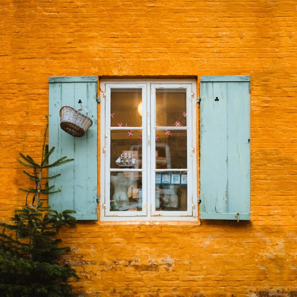 Pale blue shutters are open on a white-sill window and orange painted wall in Demark. Sororal X Denmark