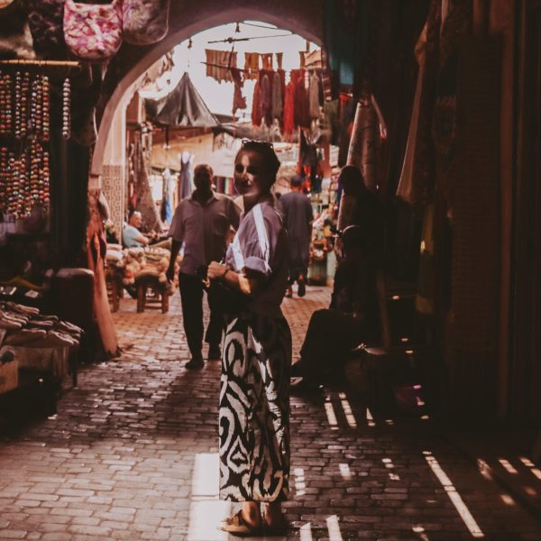 A woman standing in shadows in an alley in Morocco, wearing a long, patterned dress looks back at the camera. Sororal X Morocco