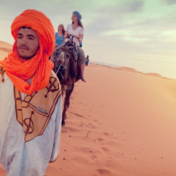 Man in a traditional outfit with a vibrant orange turban leads camels carrying tourists across vast desert sand dunes under a soft, cloudy sky.