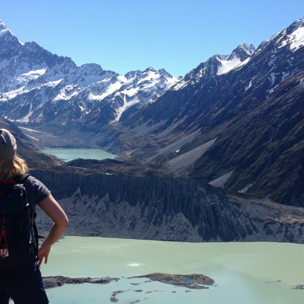 A hiker overlooks a glacial lake framed by towering, snow-capped mountains in a breathtaking New Zealand national park