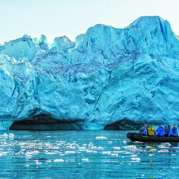 A group of people in a small inflatable boat exploring icy waters near a towering blue glacier in the Arctic - Out of the Northwest Passage