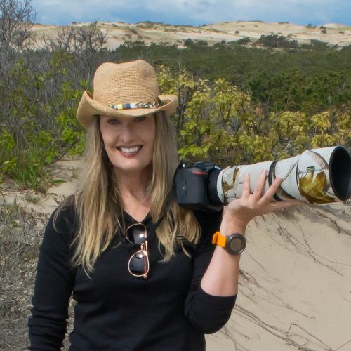 Sarah E Devlin, CEO and owner of Women in Wildlife Photography, poses for the camera wearing a straw cowboy hat and holding a long-lensed camera.