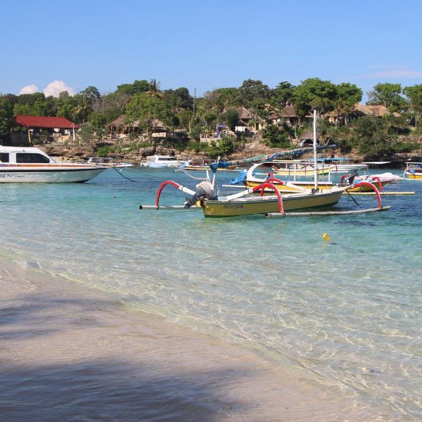 A scenic view of a beach in Bali with clear turquoise water, traditional fishing boats, and lush greenery in the background.