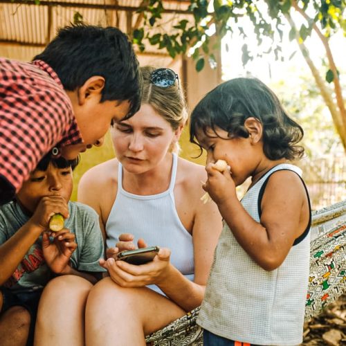 A young woman sits on a hammock surrounded by three children, all looking at a phone screen—Worldpackers volunteer abroad