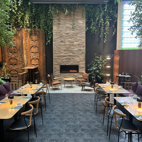 The dining area of the Amerikalinjen Boutique Hote in Oslo Norway has live edge wood tables, hanging greenery, and a modern fireplace set in a tall brick wall.