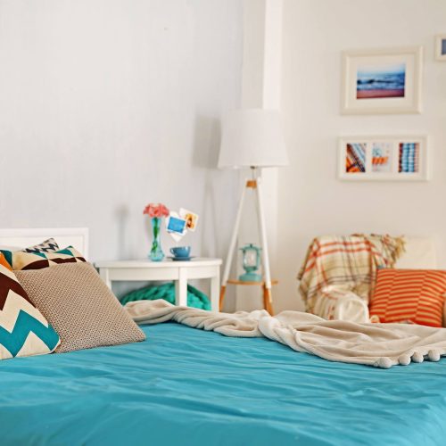 A simple bedroom with a night table, lamp and chair, and a beach-themed gallery wall. Homestay.com