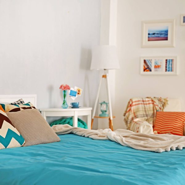 A simple bedroom with a night table, lamp and chair, and a beach-themed gallery wall. Homestay.com