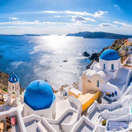 A classic panoramic view of Greece with blue-domed homes and sun gleaming on the crystal blue ocean - Collette Travel