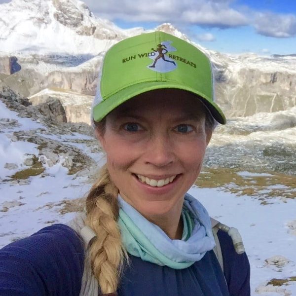 Elinor Fish, founder of Run Wild Retreats - offering mindfulness and trail running trips for women - poses for a selfie in a brand cap with a snowy mountain scene behind her.