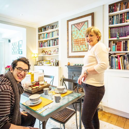 A young man sat at a kitchen table set for lunch, with an older woman standing beside the table, both smiling at the camera in a nicely decorated room with a fireplace and built in bookshelves. Homestay.com