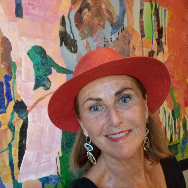 Wearing a red fedora in front of a wall mural, Nancy McGee, owner of Absolutely Southern France, smiles for a selfie.