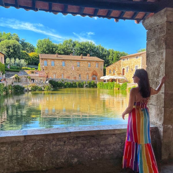 Wearing a rainbow maxi dress, Angela Macaluso stands beside a low wall over looking a river and Italian buildings for her tour Italy Awaits: A Journey for Women’s Souls