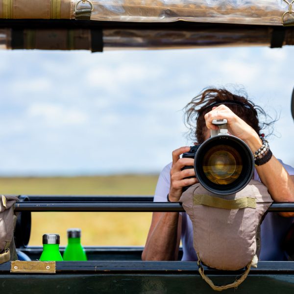 A woman with a wide-lens camera takes a photo from the back of a moving jeep on safari.