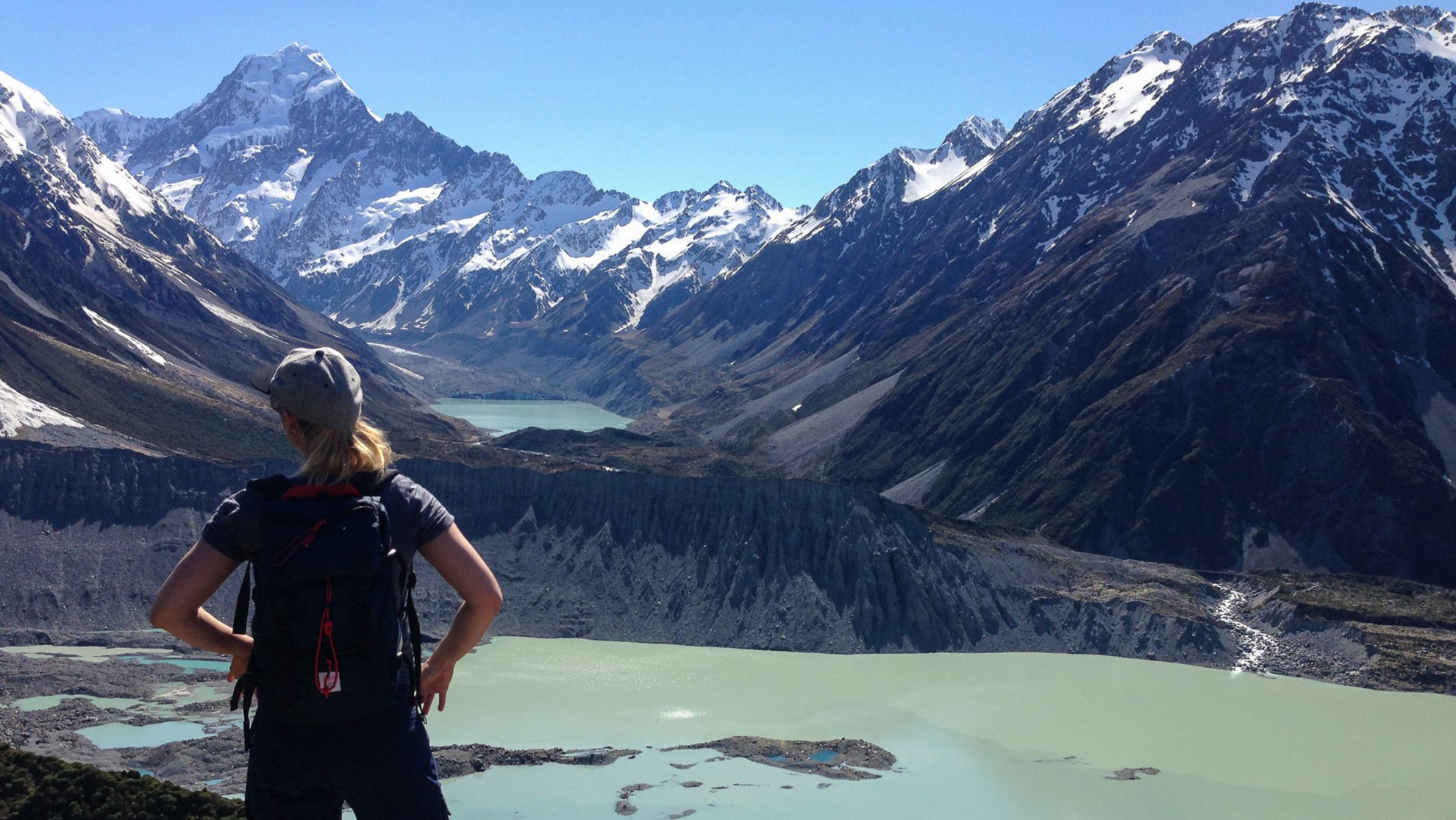 A hiker overlooks a glacial lake framed by towering, snow-capped mountains in a breathtaking New Zealand national park