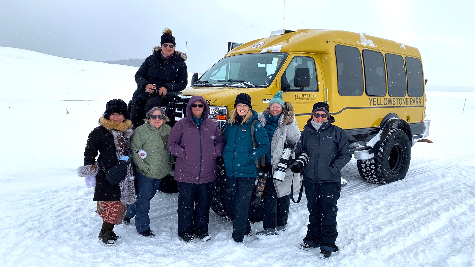 A group of women dressed in outdoor winter gear stand for a picture in front of a yellow bus on a snow-covered road as part of the Yellowstone in Winter Photography Tour by Women in Wildlife Photography