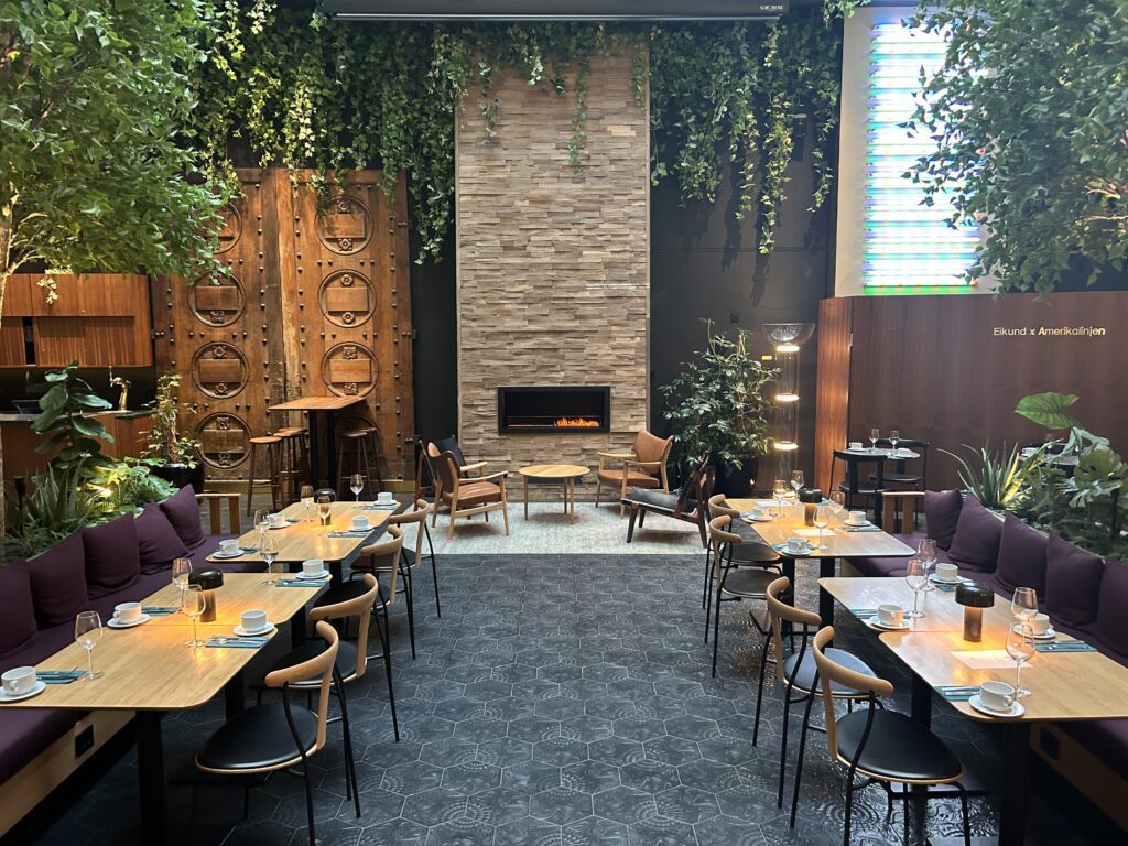 The dining area of the Amerikalinjen Boutique Hote in Oslo Norway has live edge wood tables, hanging greenery, and a modern fireplace set in a tall brick wall.