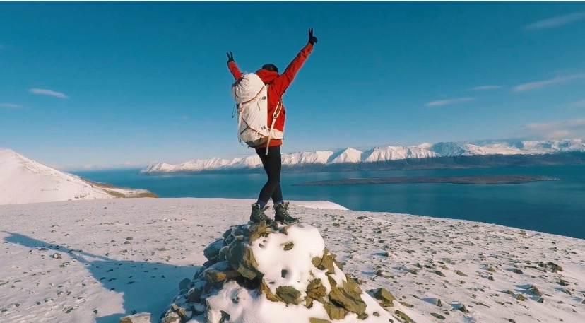 A women standing on a snowy hill with arms raised, overlooking a fjord in North Iceland.-North Iceland Yoga Adventure